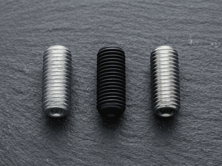 Close-up of three threaded industrial screws in silver, black, and stainless steel finishes on a dark textured surface.